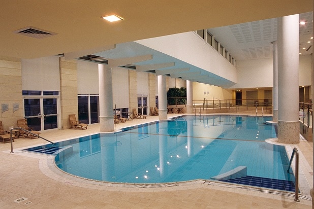 An indoor swimming pool area is shown, featuring a spacious, clear water pool surrounded by light-colored tiles. Lounge chairs are positioned around the pool, and large windows allow natural light to illuminate the space. The atmosphere appears calm and inviting.