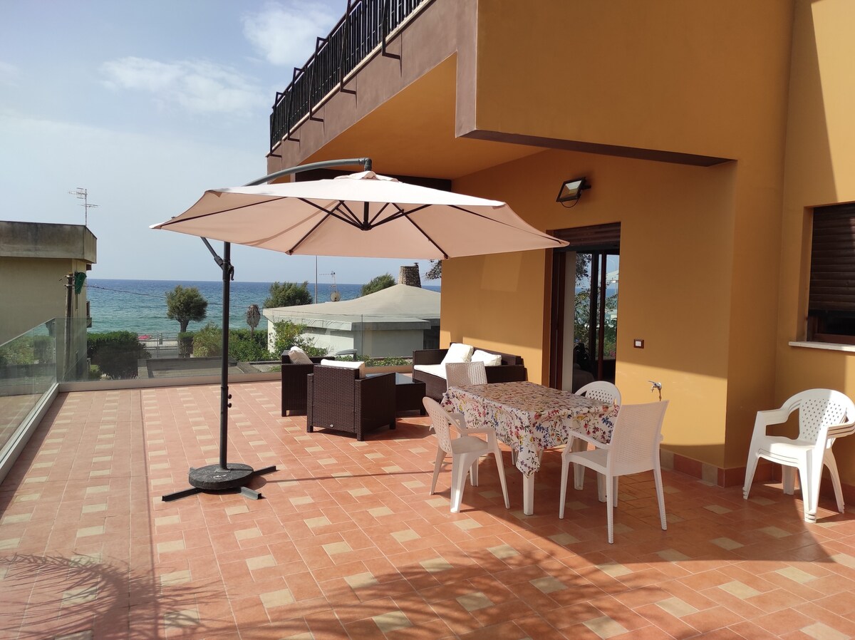 A spacious terrace is shown, featuring a dining table with an umbrella and several chairs. Comfortable seating arrangements with light-colored cushions are visible. The terrace offers a view of the sea and surrounding greenery, emphasizing the outdoor space's inviting nature.