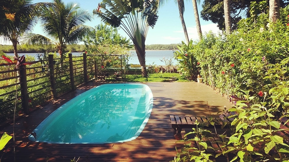 An outdoor area features a private pool surrounded by a wooden deck. Lush greenery and tropical plants frame the scene, with a view of the water visible in the background. The area offers a tranquil setting for relaxation.