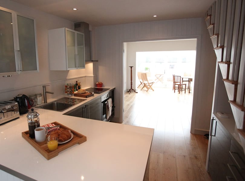An open kitchen space features modern appliances and sleek cabinetry, complemented by a clean countertop. A wooden tray displays breakfast items, including pastries and a beverage. Natural light floods the area, leading to a cozy dining space with chairs visible through the adjacent glass doors.