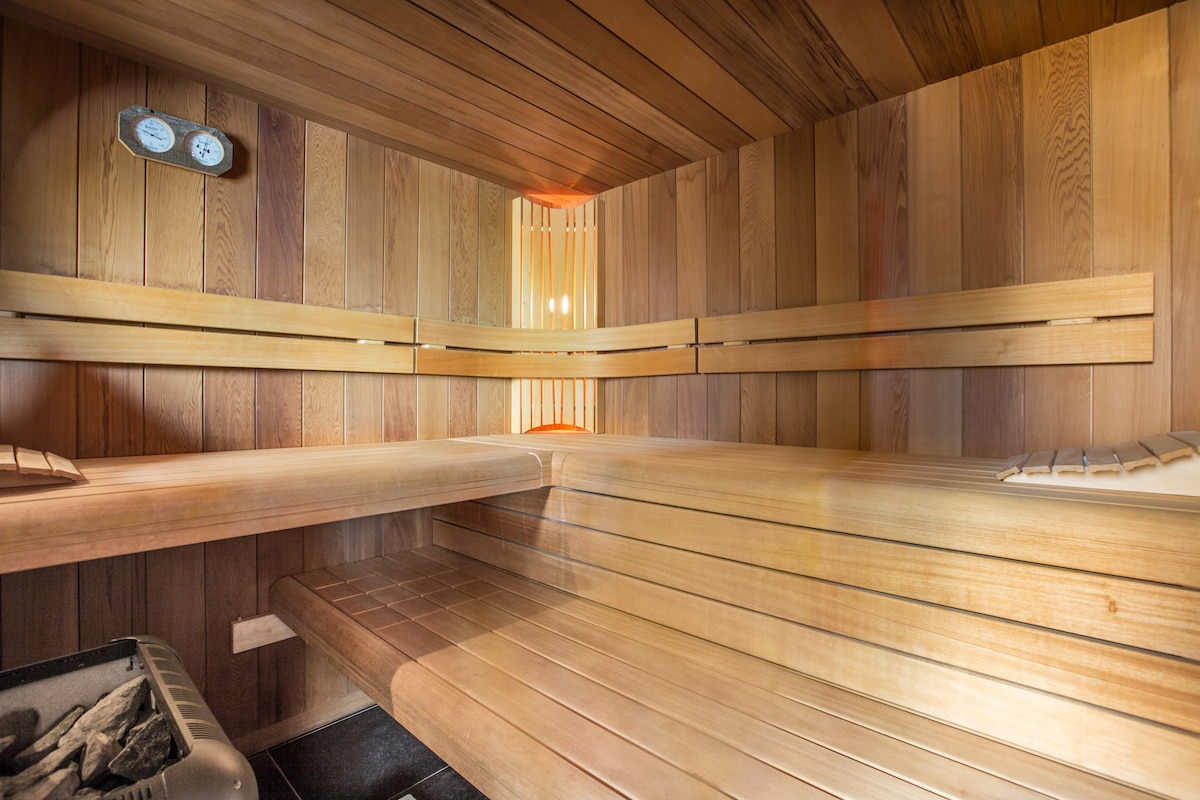 A wooden sauna is equipped with multiple benches, providing a spacious interior for relaxation. The warm wood tones create a calming environment. Wall-mounted temperature gauges are visible, indicating the sauna's functional features, while soft lighting enhances the serene atmosphere.