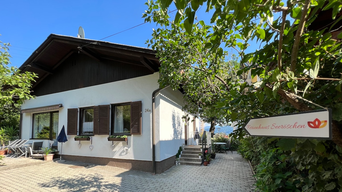 The bungalow's exterior is showcased, featuring a light-colored facade and dark wooden accents. A sign reads 'Haus Andrea Seerestaurant.' Several windows with shutters are visible, surrounded by greenery. A stone pathway leads to the entrance, flanked by comfortable outdoor seating.