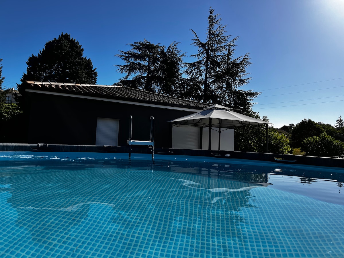 A private above-ground pool is set against a clear blue sky, surrounded by greenery. A shaded pergola provides a relaxing spot for shade, while a sleek ladder leads into the pool. The villa's modern exterior is visible in the background.