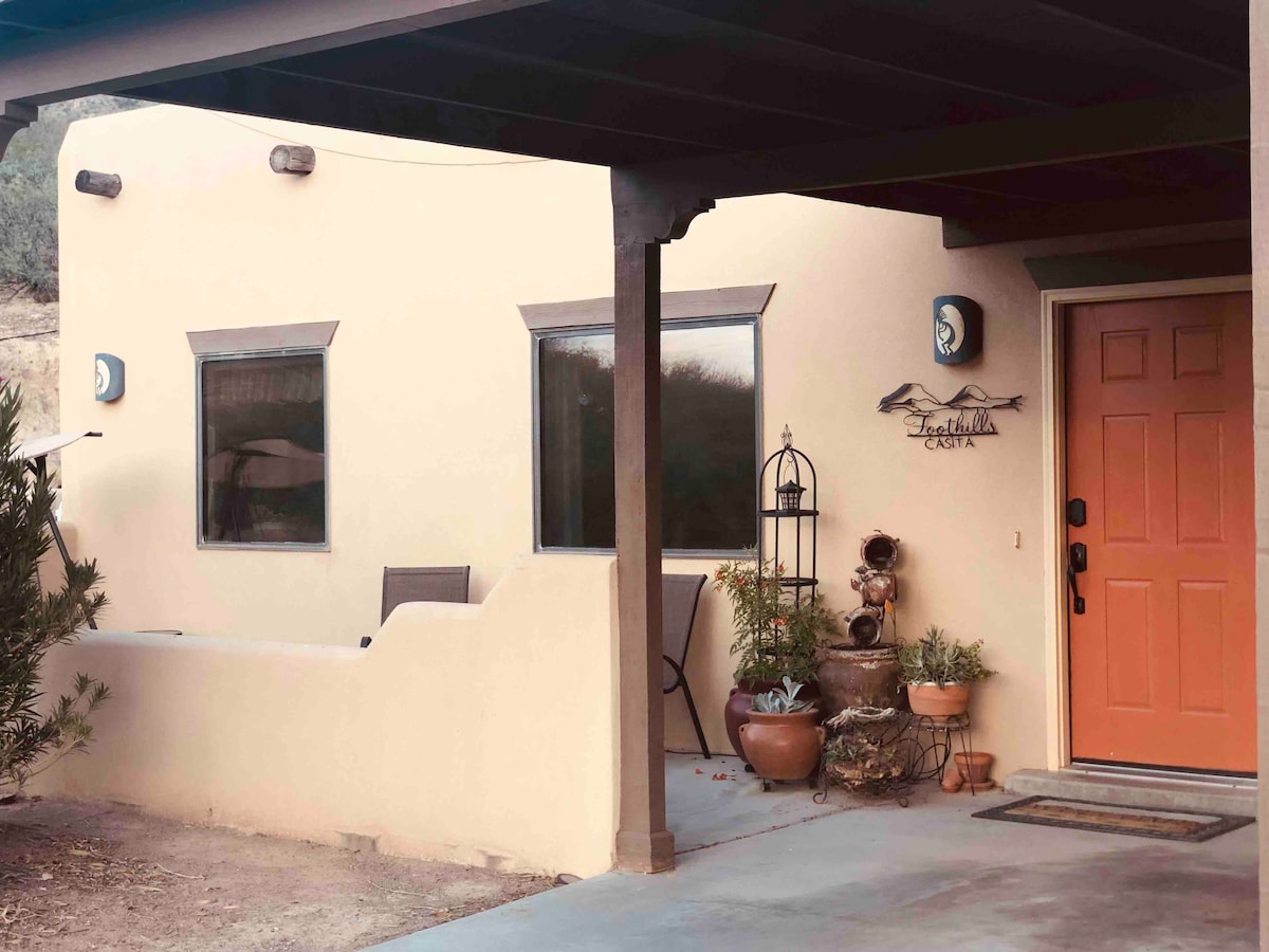 The exterior of the casita features a warm beige stucco facade with a welcoming orange door. Two windows are present, framed by black trim. Potted plants adorn the entrance, and a decorative wall piece adds character. Covered parking is visible in the foreground.