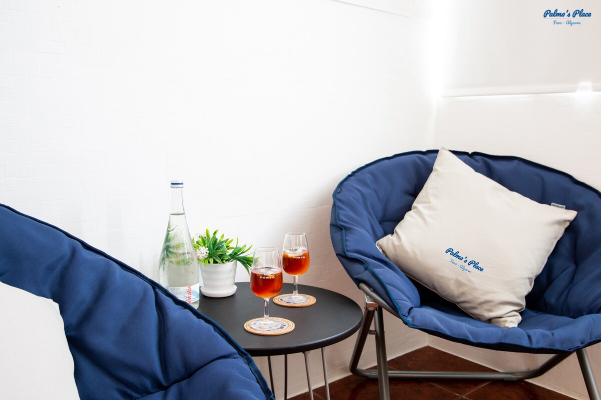 A small seating area features two blue cushioned chairs arranged around a black table. A bottle of water sits alongside two glasses filled with a red drink, complemented by a small green plant on the table. Soft light enhances the bright, inviting atmosphere.