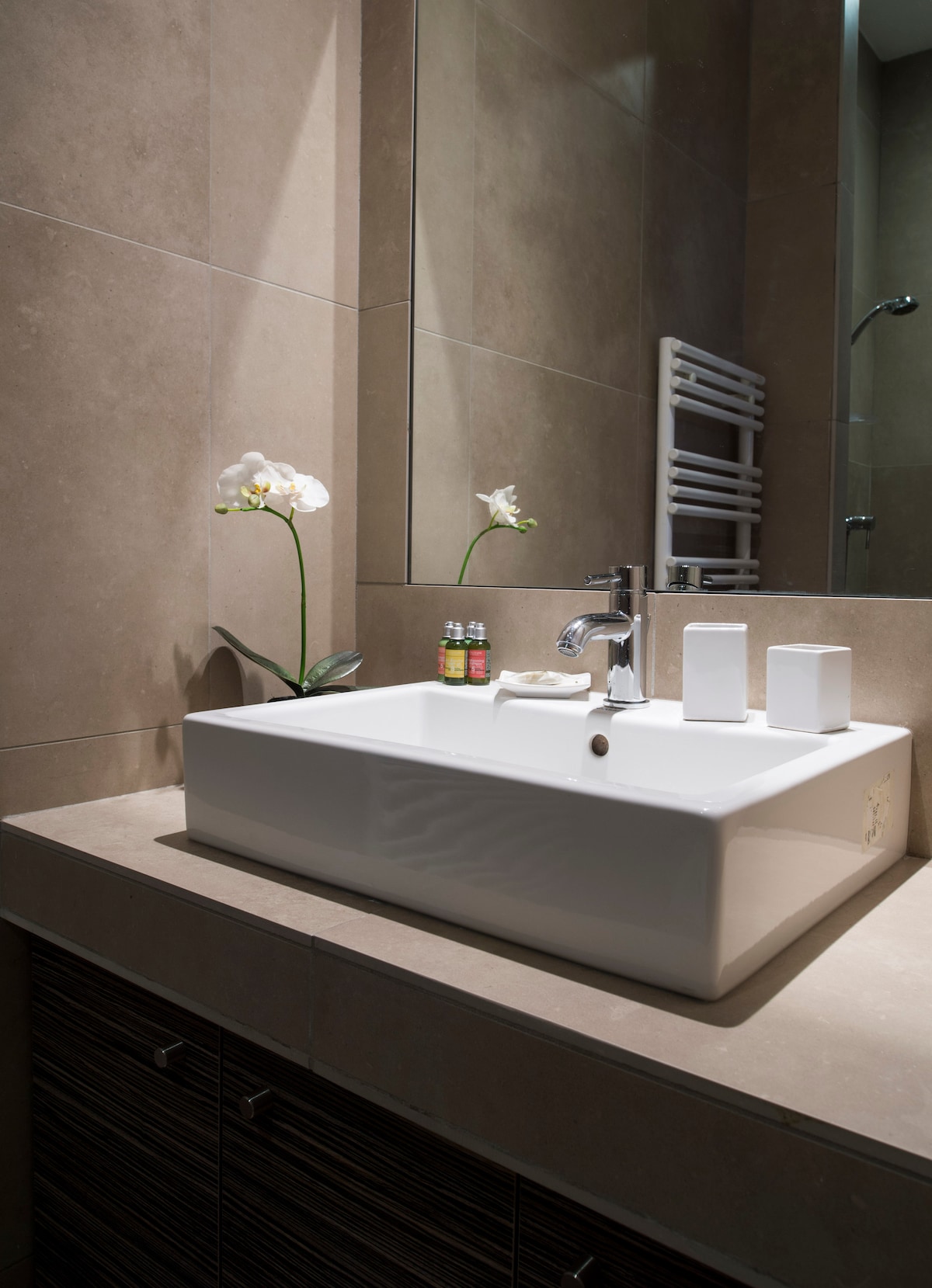 A contemporary bathroom features a white sink integrated into a stone countertop. A mirror reflects the soft lighting, while two elegant orchid plants add a touch of greenery. Amenities include toiletries placed beside the sink and a towel rack on the wall.