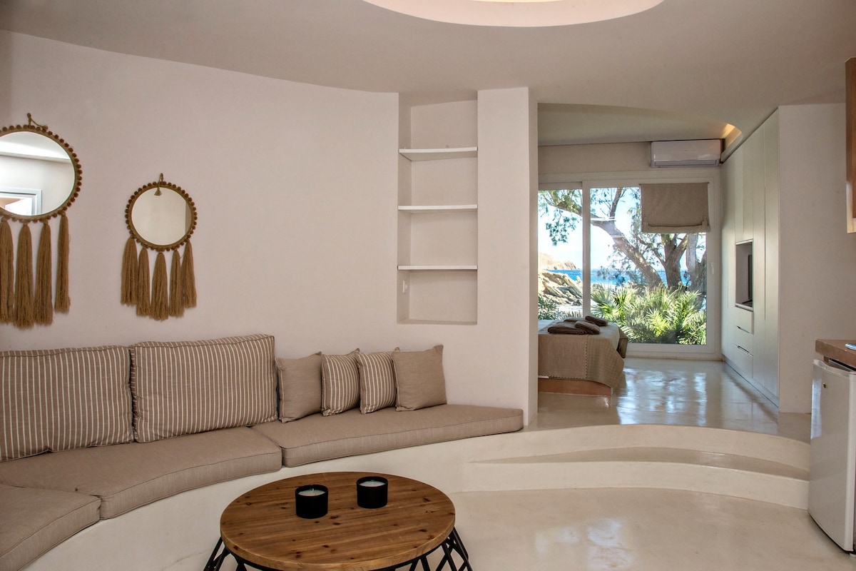 A spacious living area features a comfortable, curved sofa with neutral-toned cushions. A round wooden coffee table sits in the center. Large glass doors provide a side view of the garden and sea, allowing natural light to brighten the space.