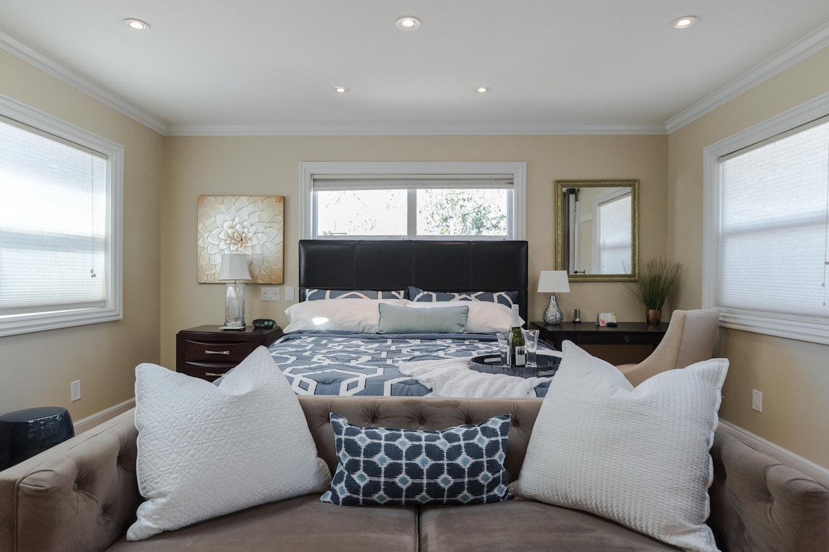 A spacious bedroom is depicted, featuring a large bed with a patterned cover and decorative pillows. Natural light enters through two windows, illuminating the room's light-colored walls and furnishings. A stylish lamp and a mirror are visible on either side of the bed, along with a cozy seating area in the foreground.