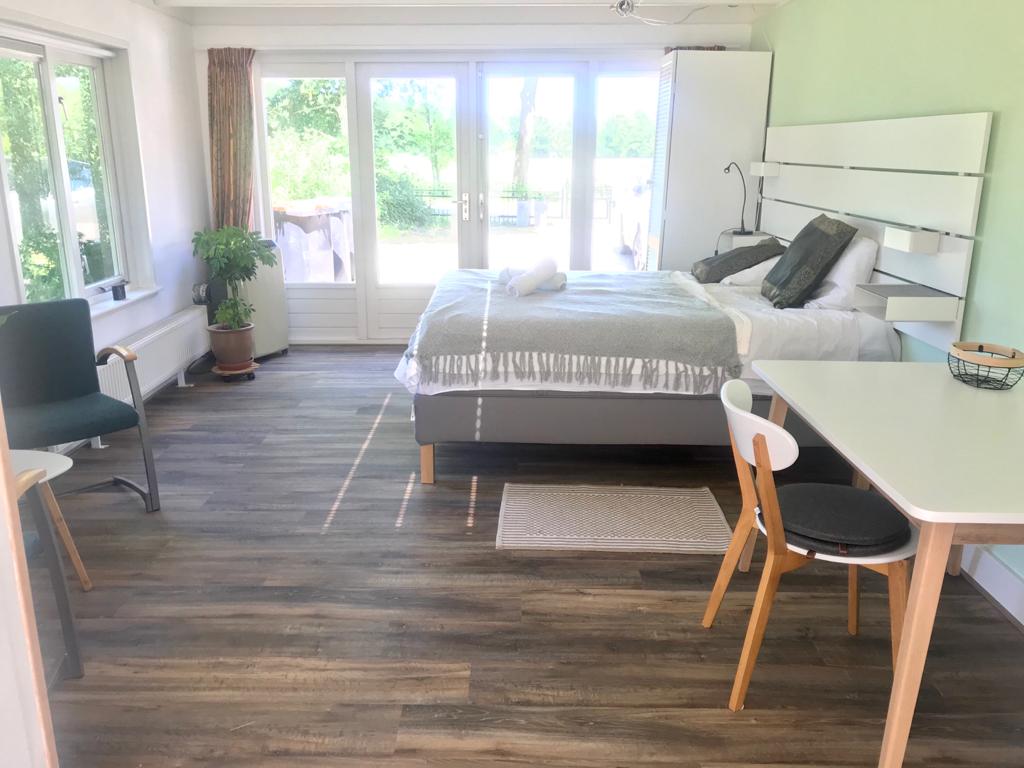 A bright and airy living space features a spacious bed with soft linens and decorative pillows. Large glass doors allow natural light to enter, leading to a view of greenery outside. A small dining table and two chairs are arranged near the window.