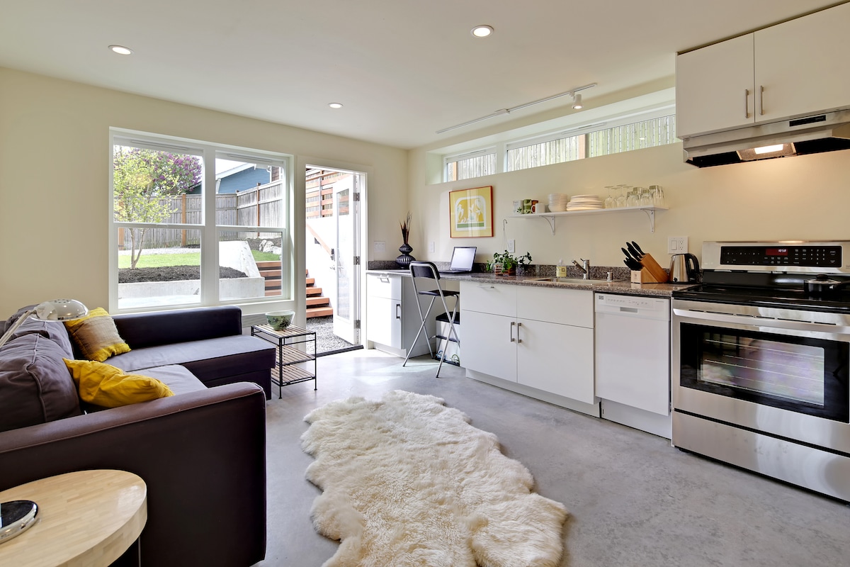 A modern kitchen area features sleek white cabinets and stainless steel appliances, including a stove and oven. A spacious countertop is equipped with kitchen essentials. Natural light fills the space through large windows, while a cozy seating area is furnished with a sofa and a decorative area rug.
