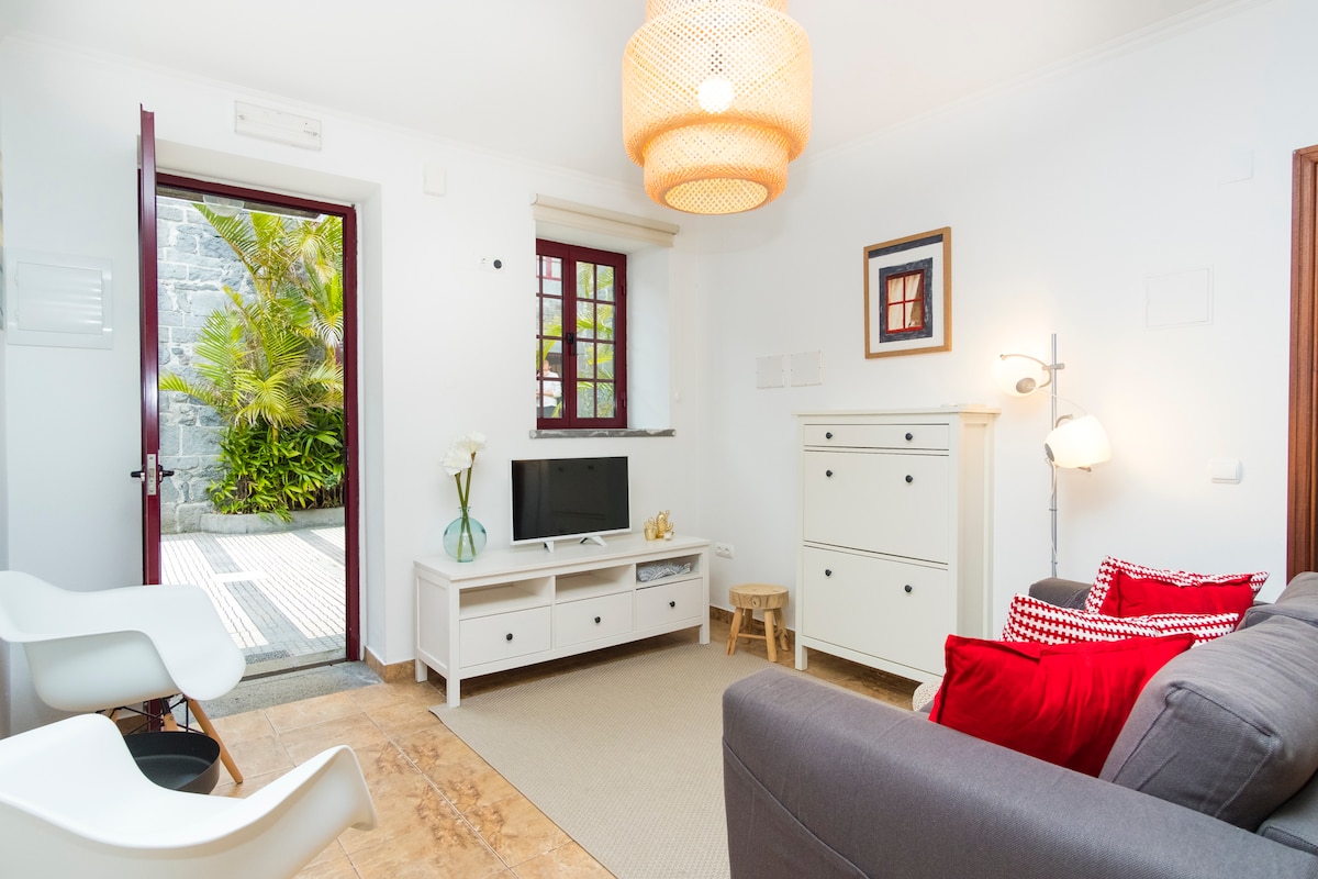 A welcoming living area features a comfortable gray sofa adorned with red cushions. A white entertainment unit houses a flat-screen TV. Light enters through a door leading to a garden, while a woven pendant light hangs overhead, complementing the warm tile floor.