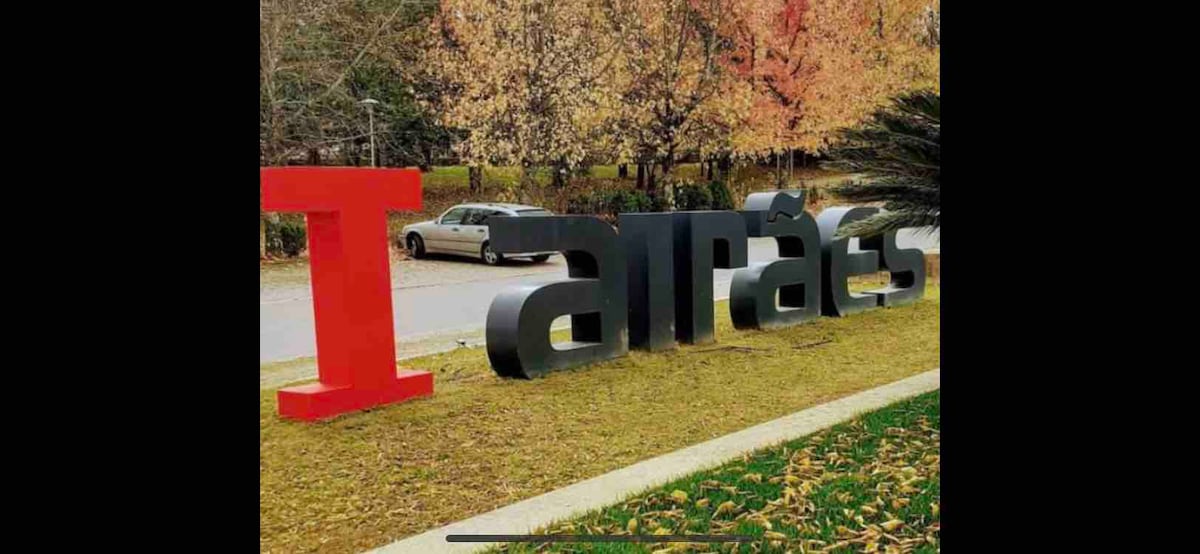 A large sign displaying the phrase 'I airães' is prominently set in a grassy area, with colorful autumn leaves scattered on the ground. The background features trees with vibrant foliage, and a car is parked nearby.