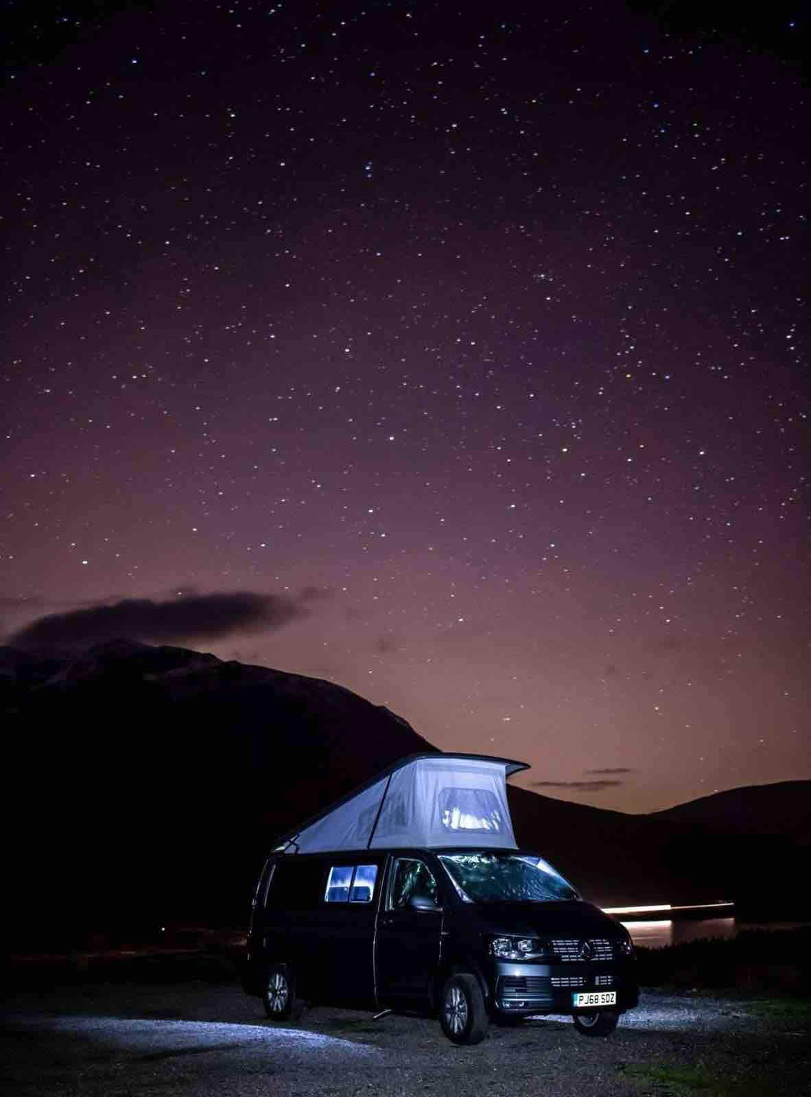 A Volkswagen campervan is parked under a starry night sky, with the iconic pop-top roof raised. The surrounding landscape features dark silhouettes of mountains, and a soft glow emanates from nearby vehicles, highlighting the peaceful camping atmosphere.