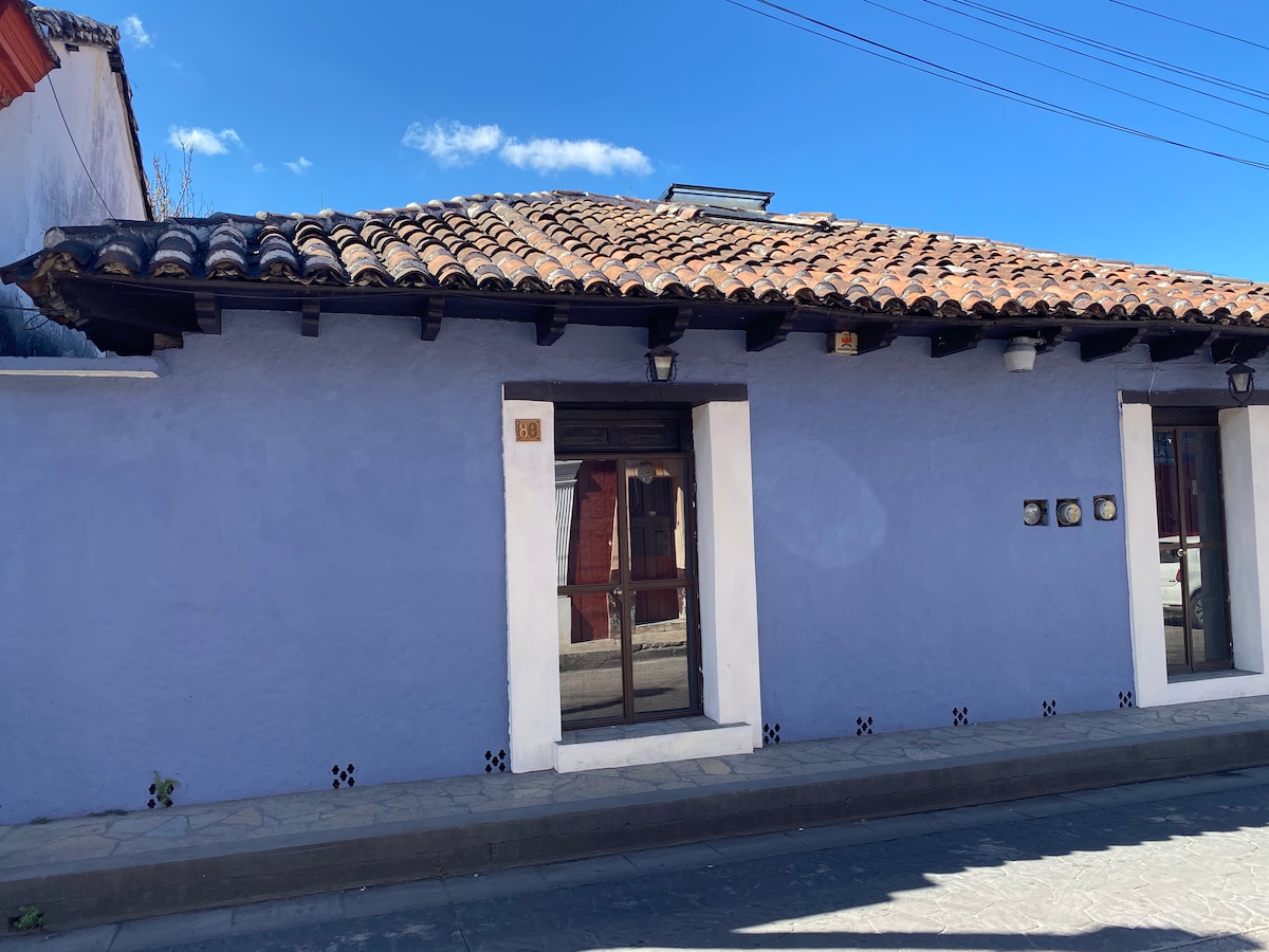 A charming exterior of a lavender building is displayed, featuring traditional tile roofing and wooden beams. The entrance is framed by a large window with wooden accents, providing natural light to the interior. Soft sunlight highlights the textured wall surface.