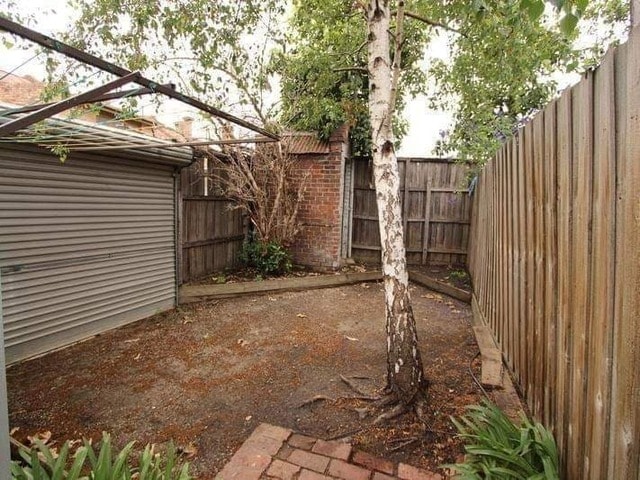 An enclosed outdoor space is presented with gravel ground and a small tree situated near a wooden fence. The area features a shed along one side, surrounded by low-maintenance vegetation, providing a private setting ideal for relaxation.
