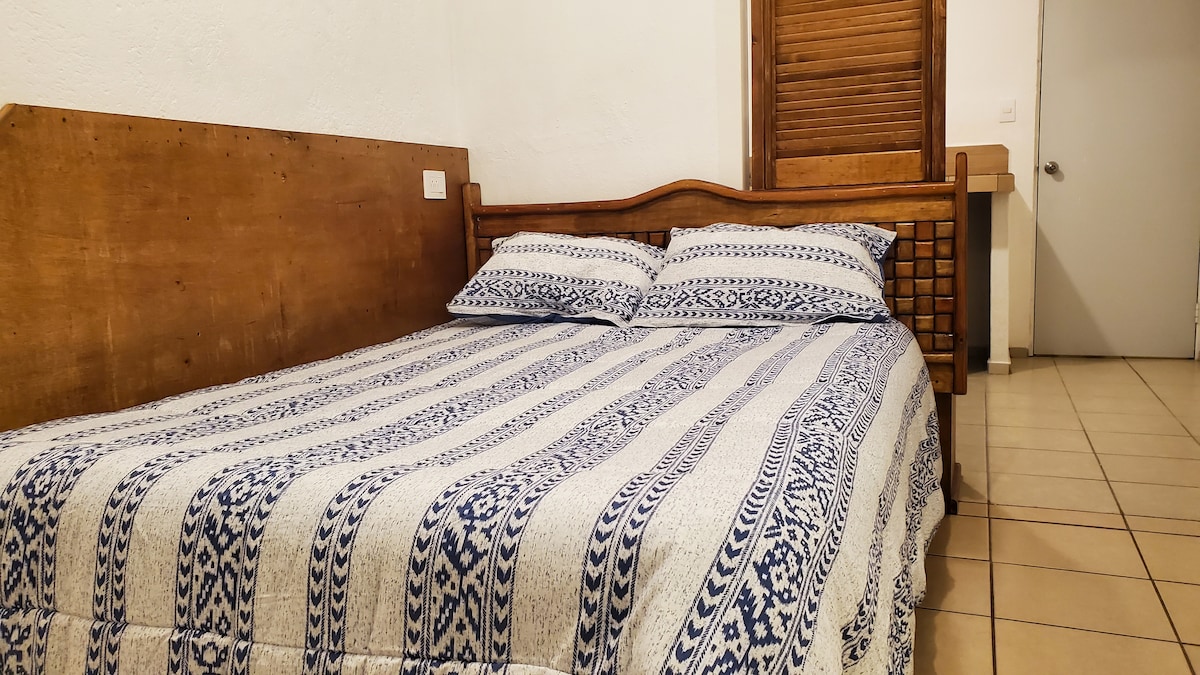 A spacious bed is adorned with a patterned blanket featuring blue and white tones. Pillows are arranged neatly at the head of the bed. A wooden headboard adds a rustic touch, with a light-colored wall and a door visible in the background.