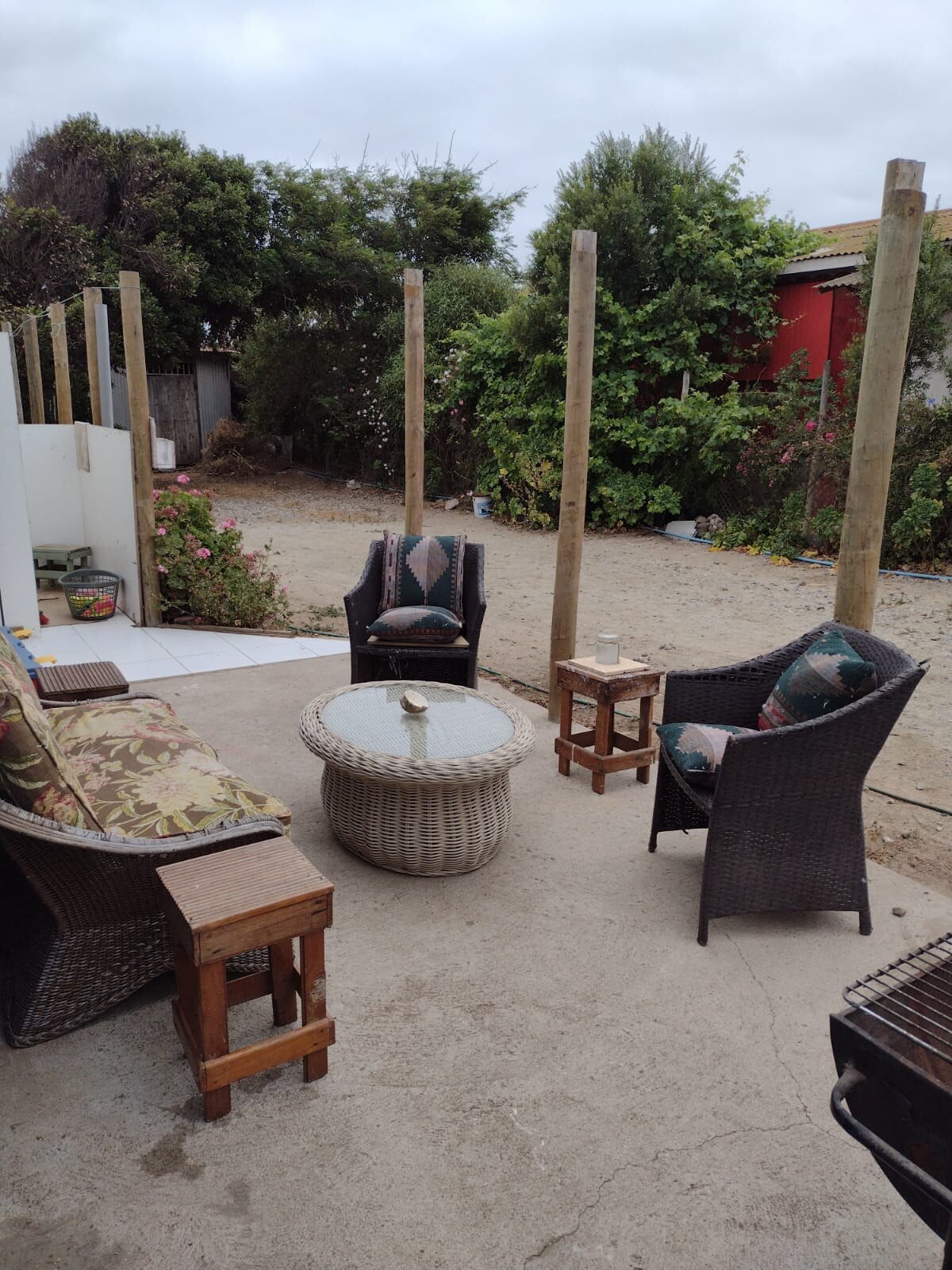 A cozy outdoor seating area features comfortable wicker chairs with patterned cushions arranged around a circular table. The space is surrounded by natural greenery and a gravel ground, creating a serene environment for outdoor relaxation.