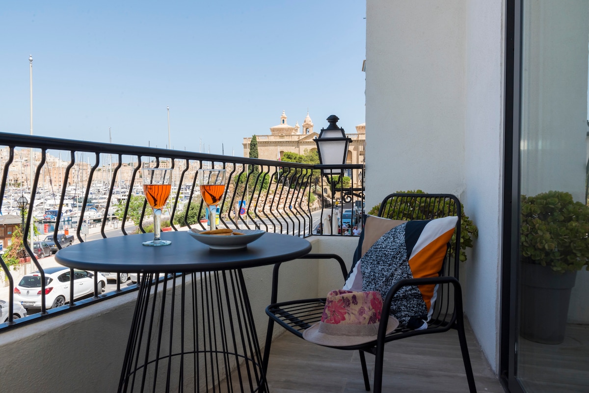 A balcony space features a round table set with two glasses and a plate. A black wire chair is positioned beside the table. The view showcases a marina with boats, complemented by historic architecture in the background.