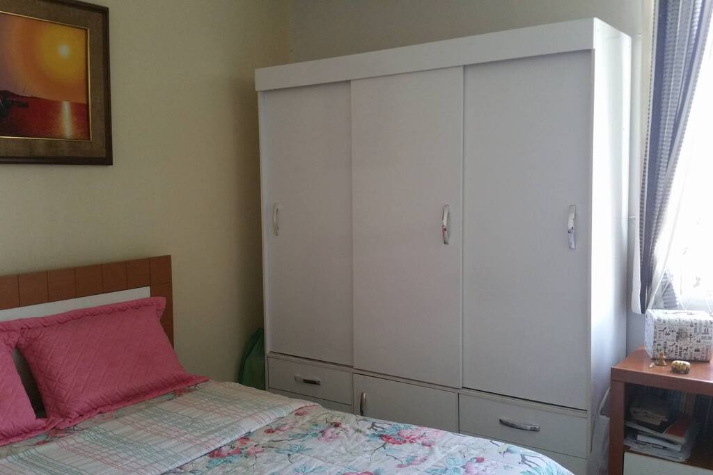 Quarto com cama grande (casal), guarda-roupa, ventilador e uma pequena estante para livros.
