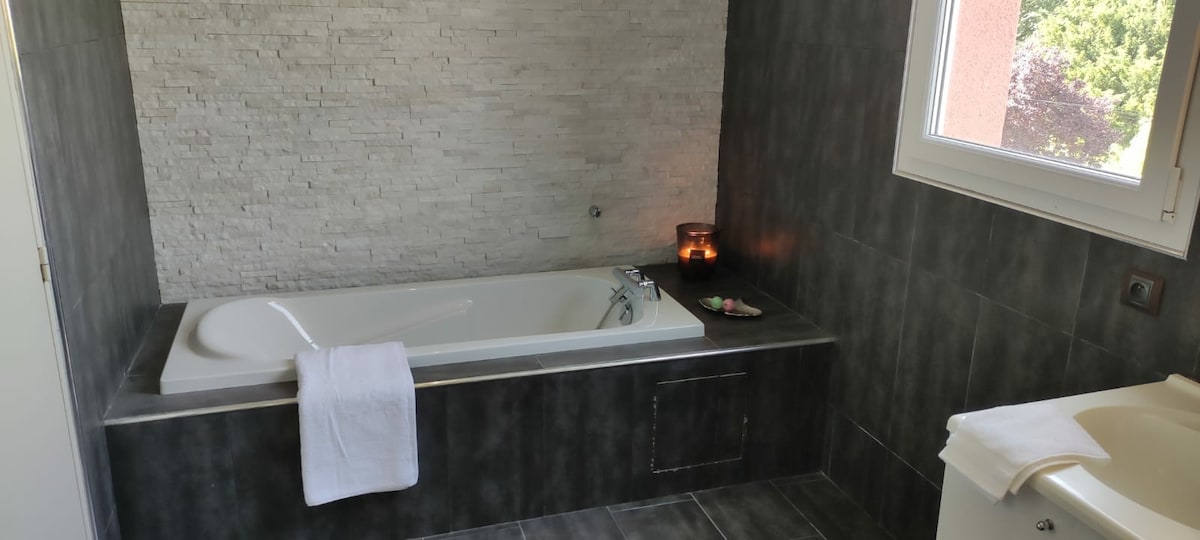 A modern bathroom features a spacious bathtub with a textured stone wall in the background. A small tray with candles and toiletries is positioned nearby, while natural light streams in through a window, illuminating the clean lines and neutral tones of the space.