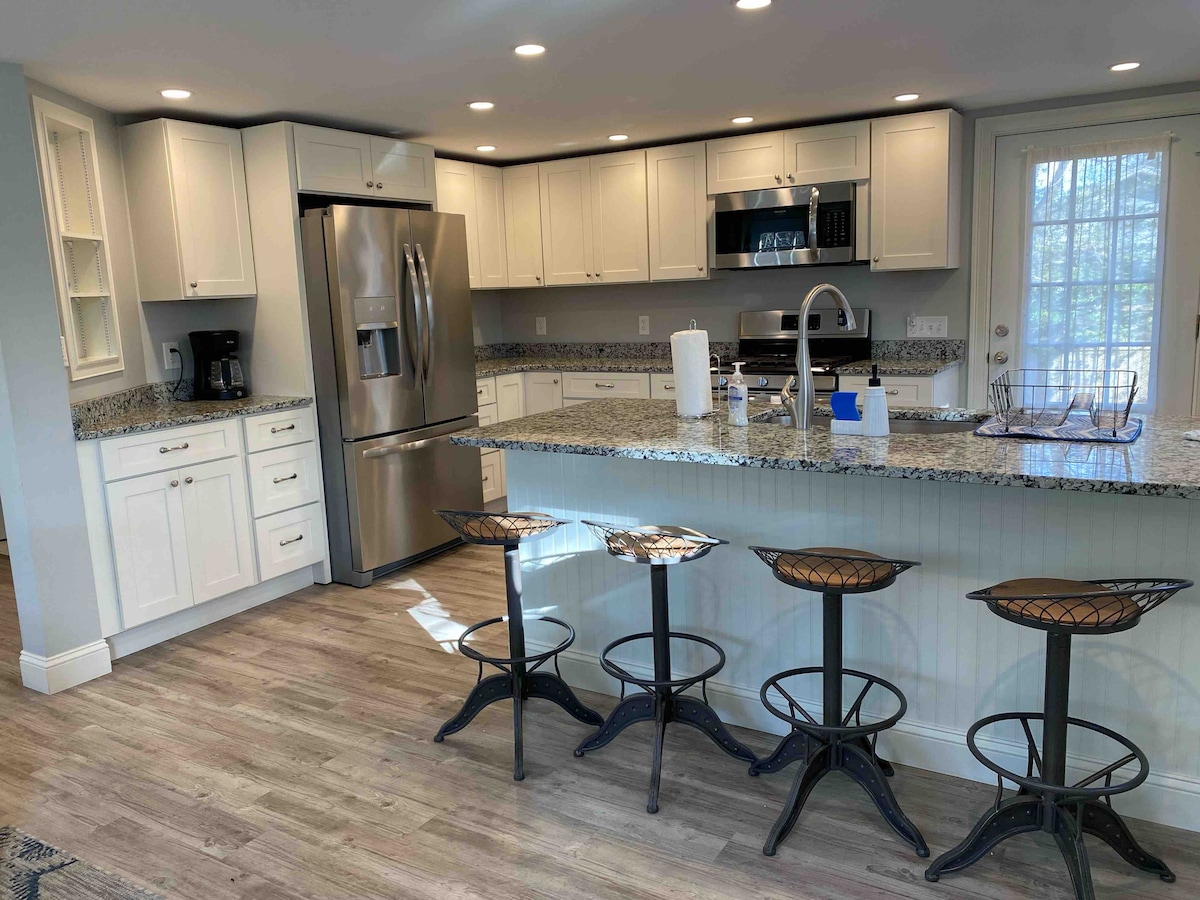 A modern kitchen features white cabinetry and granite countertops, complemented by stainless steel appliances. Bar stools are positioned at the kitchen island, and natural light filters through the nearby door, illuminating the warm wood flooring.