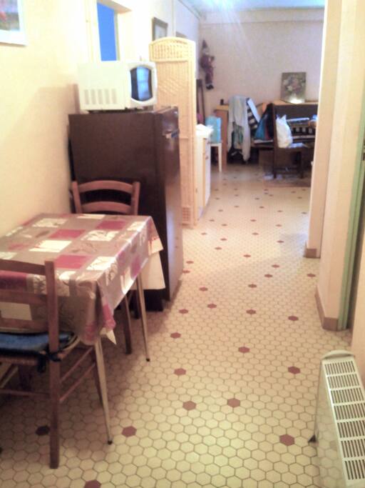 L'espace cuisine avec frigo, micro-onde, table et ses chaises, gazinière. Lavabo dans la pièce à côté.
