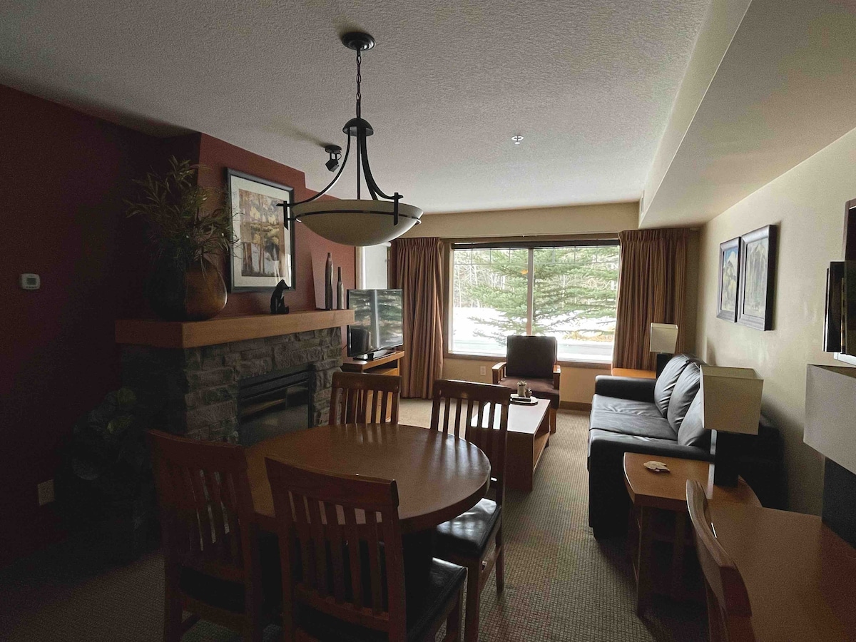 A welcoming living area features a stone fireplace and large windows with views of the outdoors. A dining table with chairs occupies a corner, while a cozy seating arrangement includes a pull-out couch and a side chair. Warm-toned walls enhance the inviting ambiance.