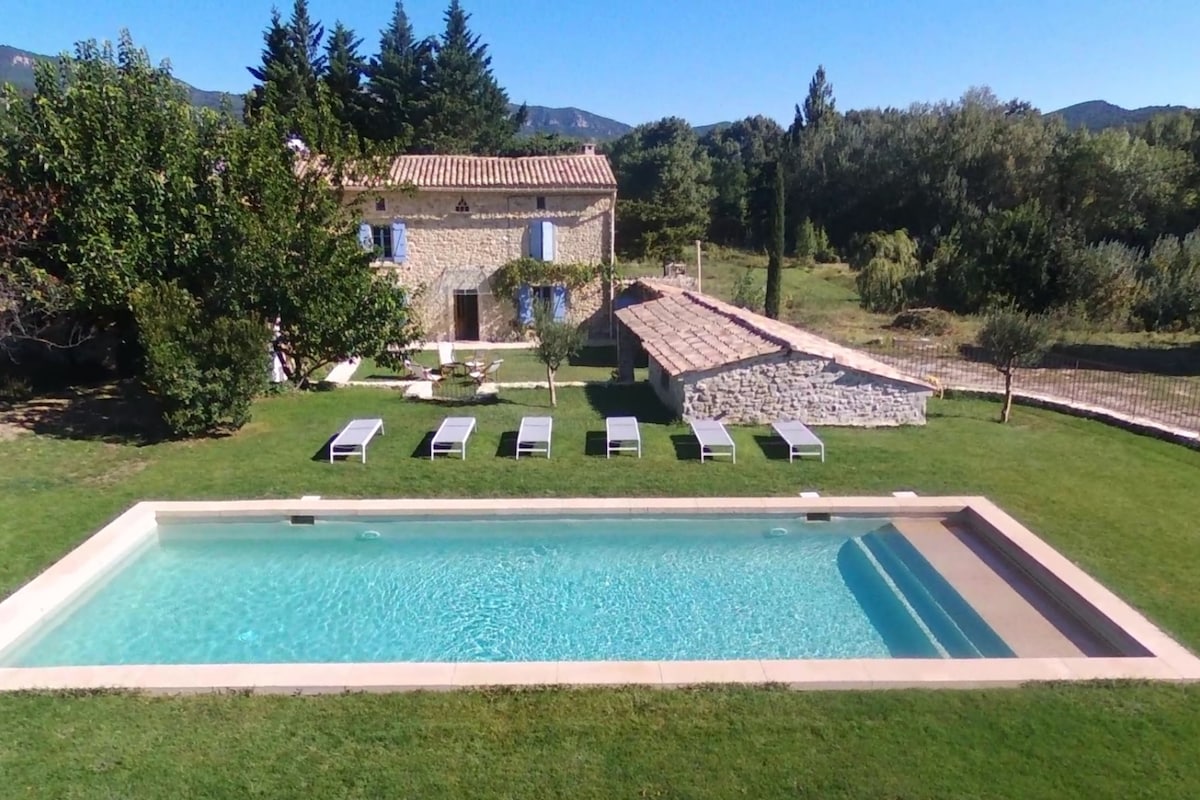 A serene outdoor area features a spacious pool surrounded by well-maintained grass and lined with six sun loungers. A rustic stone house is visible in the background, complemented by trees and scenic hills, creating a tranquil environment.