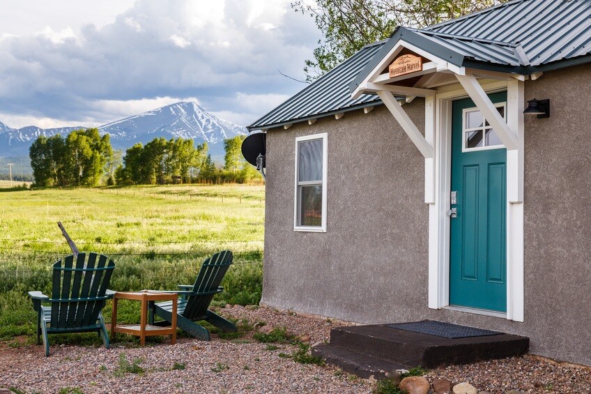 A cottage entrance is framed by a teal door, complemented by a small wooden step. Two green Adirondack chairs and a table are positioned on gravel, facing an expansive landscape of green fields and distant mountains under a partly cloudy sky.
