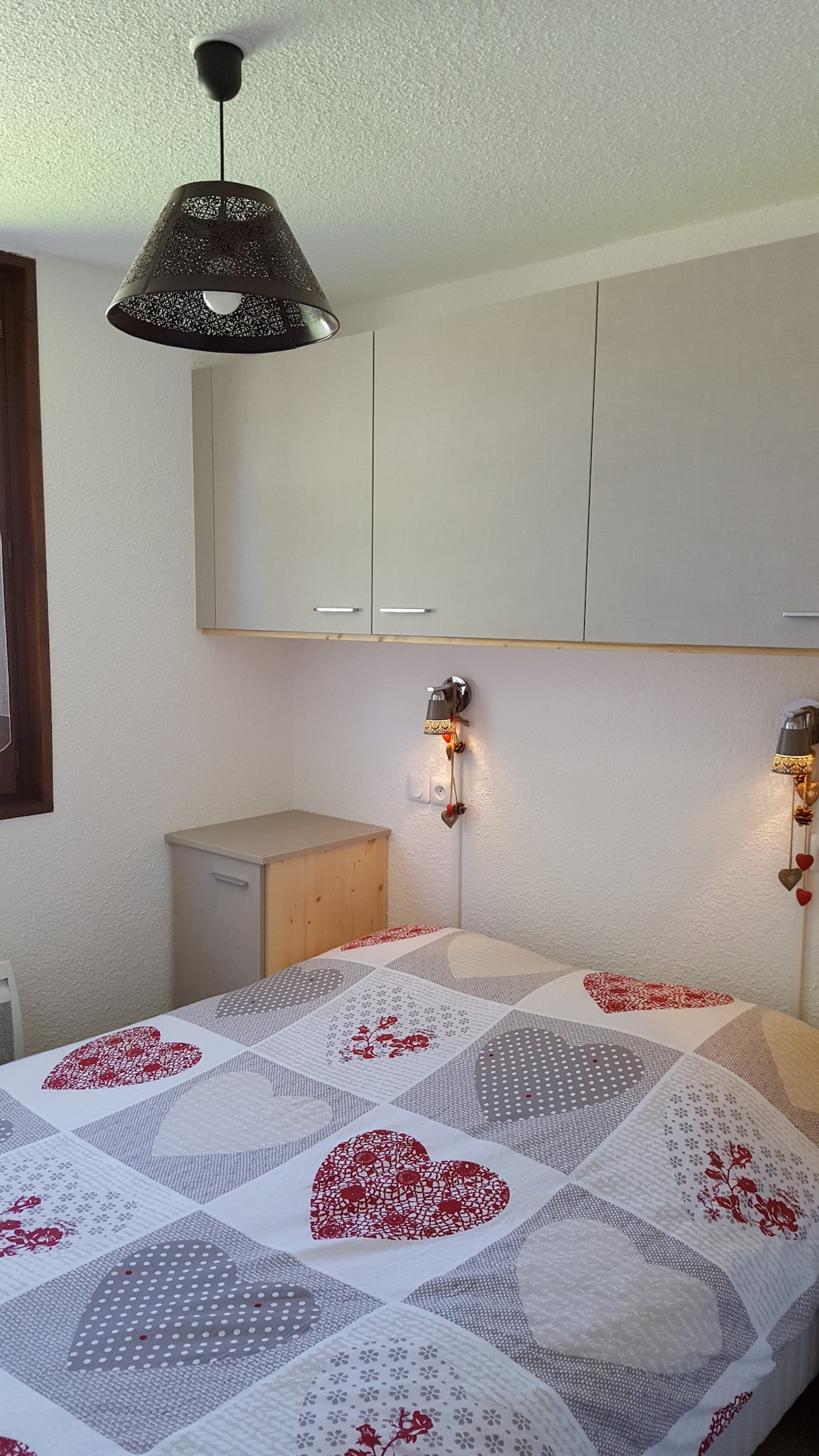 A sleeping area features warm lighting from two bedside fixtures and a patterned quilt that covers a spacious bed. Above the bed, cabinets provide additional storage, while a nightstand offers convenience. Light enters through a window dressed with subtle curtains.