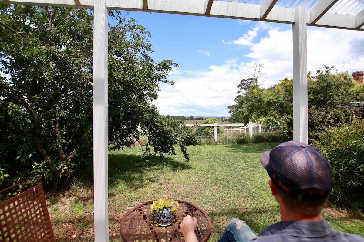 A patio area showcases a small round table with a potted plant placed on top. A seated individual is seen gazing across the green lawn toward an expansive view of fields and distant trees under a partly cloudy sky.