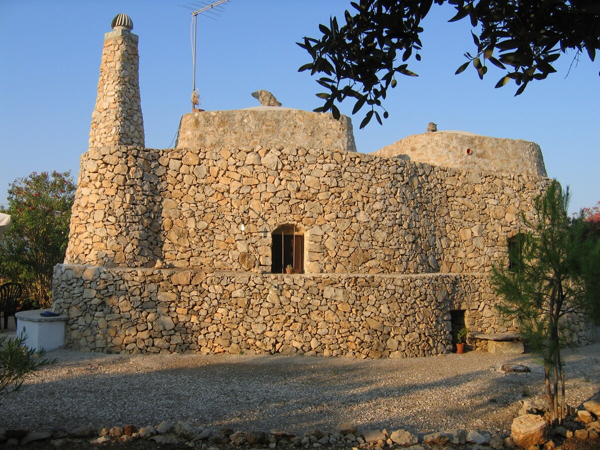 A traditional stone trullo with a conical roof is surrounded by a gravel path. The structure features smooth, unbroken stone walls and two arched windows. A chimney sits prominently on top, indicating the presence of a fireplace inside.