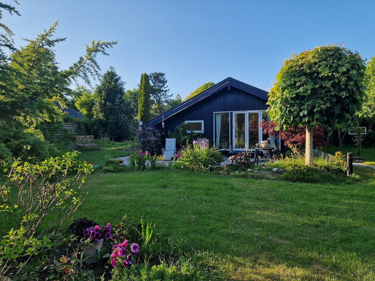A charming summer house is set among lush greenery, featuring a dark exterior and large glass doors that invite natural light. Colorful flowers bloom in the foreground, while well-maintained trees provide shade in the spacious garden visible in the background.