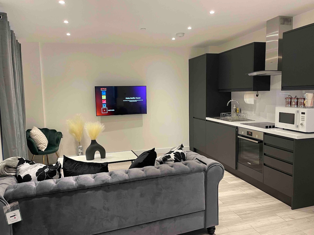 An open living space features a plush gray sofa paired with decorative pillows. A modern kitchen is equipped with sleek cabinetry, an oven, and a microwave. A wall-mounted television is visible, showcasing a streaming interface, while soft lighting enhances the inviting ambiance.