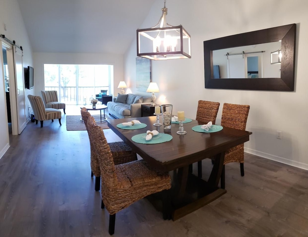 An open dining area is seen with a rectangular dark wooden table surrounded by four woven chairs, set with decorative plates and glasses. A spacious living room area is visible in the background, featuring a light-colored sofa and large windows letting in natural light.