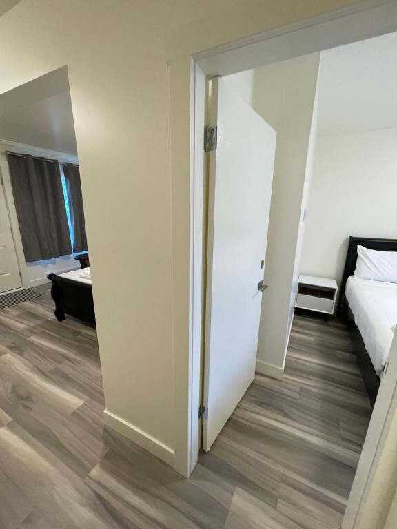 A well-lit hallway is shown, with a closed door on the left and an open entryway to a bedroom on the right. The flooring is a light wood tone, and the walls are painted in a neutral color. Grey curtains frame a nearby window.