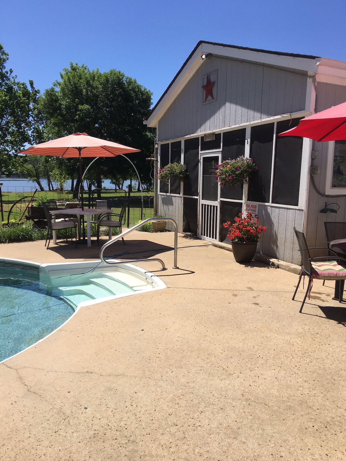 A patio area is visible beside a clear pool, featuring lounge chairs and shaded tables under colorful umbrellas. Planters with vibrant flowers accentuate the space. The screened-in building stands in the background, while lush greenery and a calm water view complete the setting.