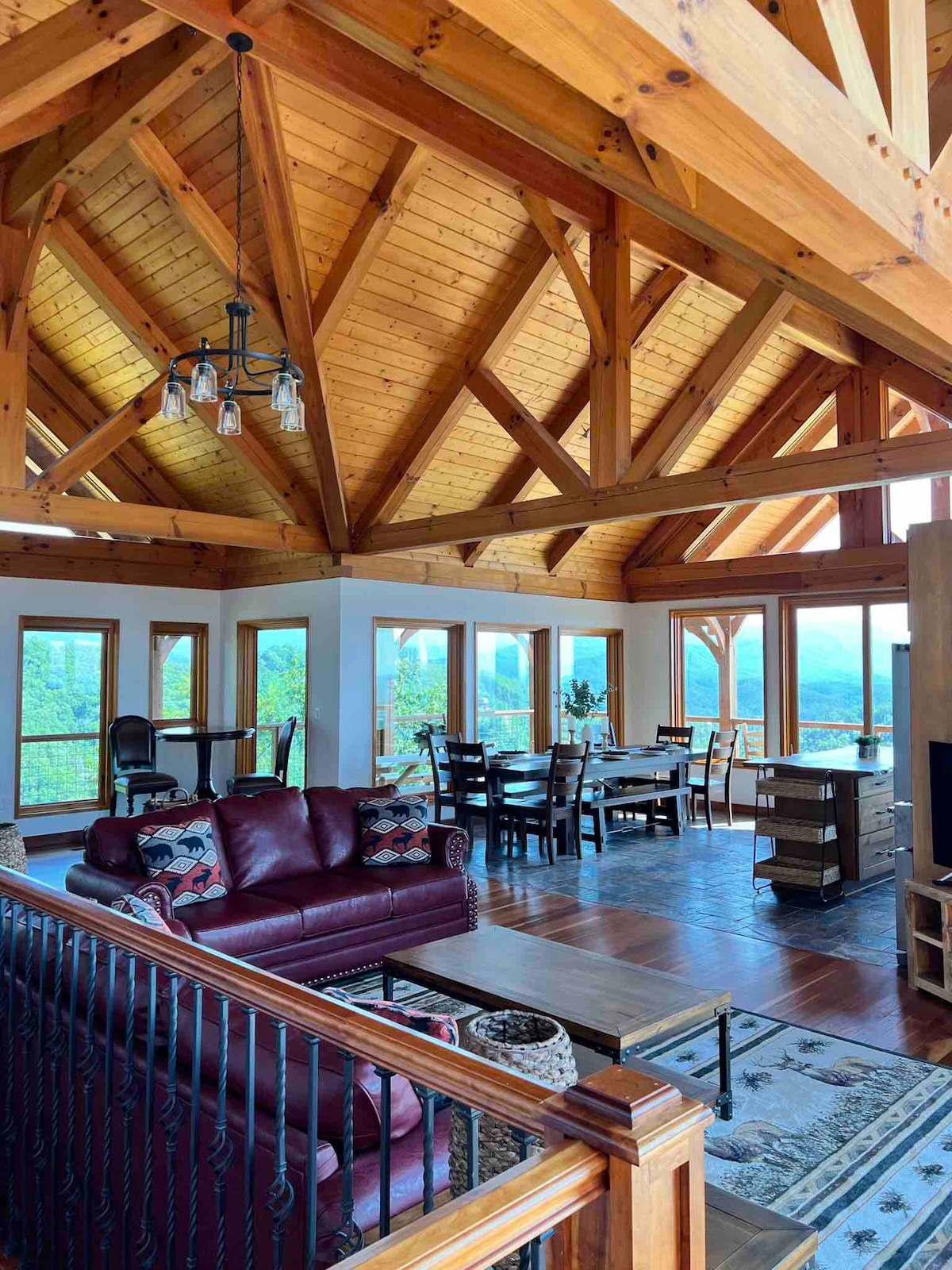 An open-plan living space is highlighted by a vaulted wooden ceiling and large windows, providing views of the surrounding mountains. A cozy red sofa with patterned pillows is positioned near a wooden coffee table, while a dining area is visible in the background.