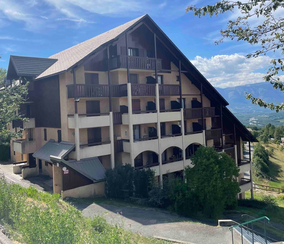 A multi-level building is situated among greenery, featuring multiple balconies that overlook a scenic valley. The structure showcases a blend of wood and stone elements, with arched windows and a sloping roof, creating an inviting presence in the landscape.