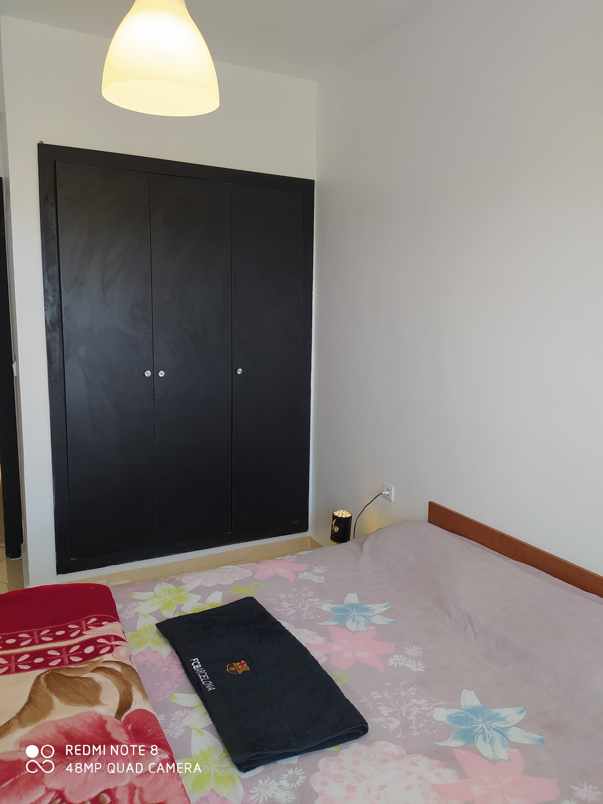 A neatly arranged bedroom features a spacious bed with a floral-patterned coverlet. A dark wardrobe with two doors occupies one wall, while a soft light hangs from the ceiling. A folded towel is placed at the foot of the bed, adding a touch of comfort.