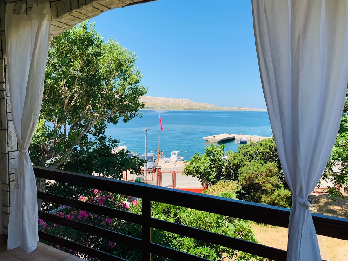 A view from a balcony showcases a calm body of water, framed by lush greenery and blooming flowers. The distant shoreline features a small harbor with boats, while a clear sky enhances the serene coastal atmosphere.