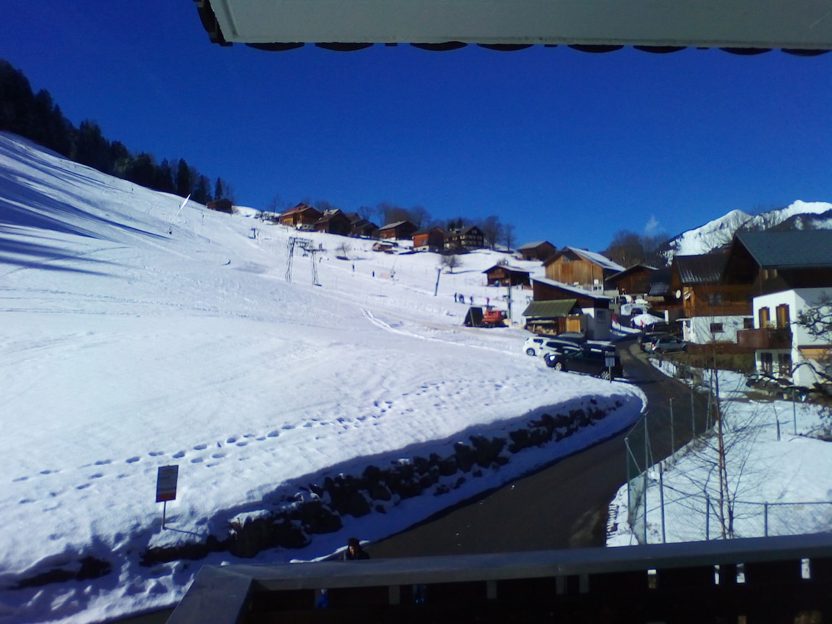 A snow-covered landscape showcases a collection of charming wooden houses nestled in the hillside. The clear blue sky contrasts the white snow, while a winding road leads through the scenery, displaying tire tracks and signs of winter activity.