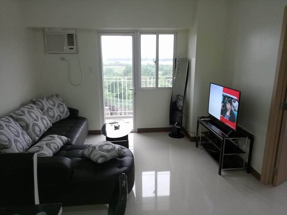 A cozy living area features a dark leather sofa adorned with circular patterned cushions. A sleek television is positioned on a glass stand, while a small round coffee table sits nearby. Natural light enters through large windows, offering views of the surroundings.