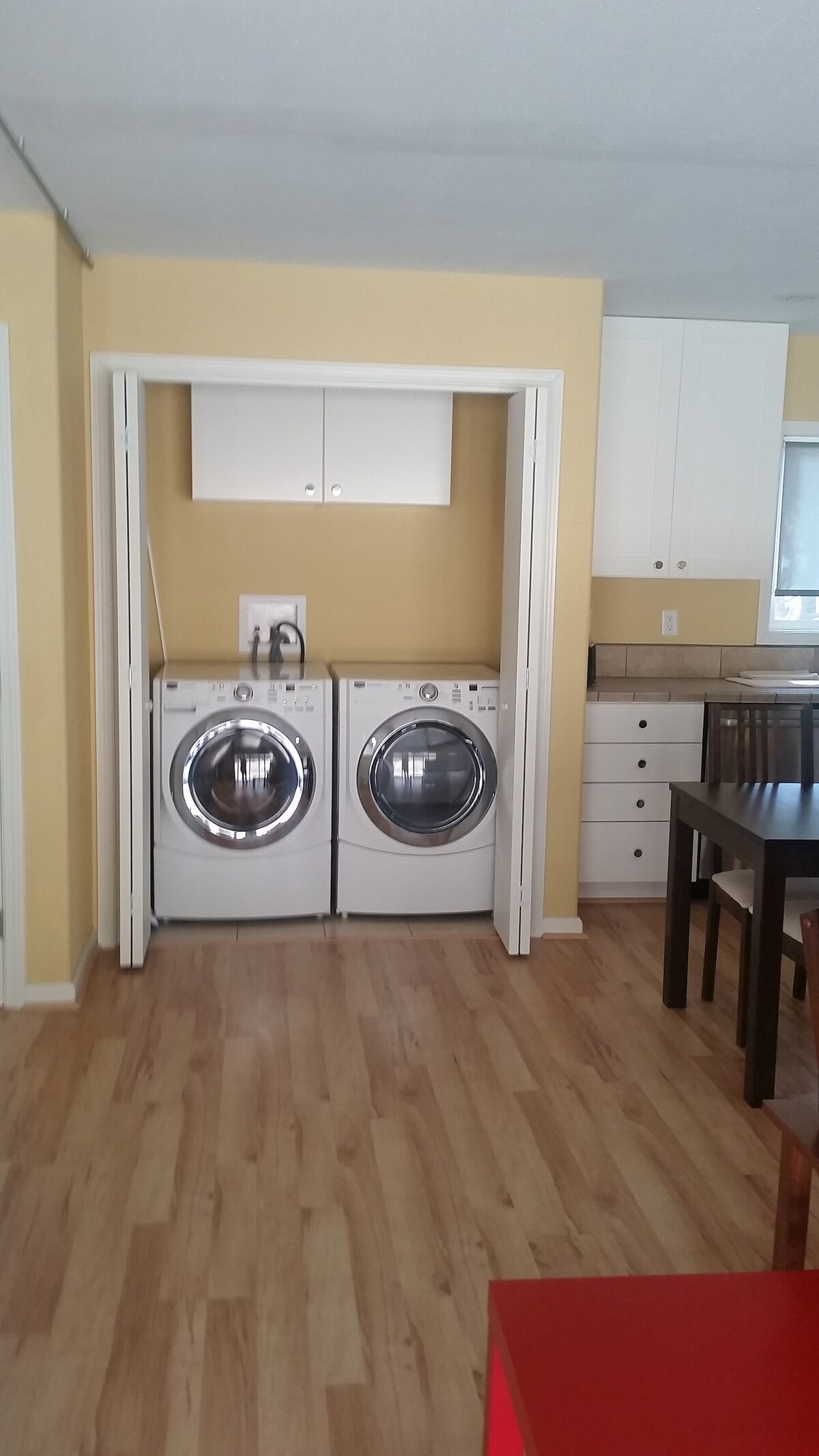 A neat laundry area is integrated into the kitchen space, featuring a set of front-loading washing machines beneath a countertop. Above the machines, cabinetry provides storage, and a small sink is visible. Light yellow walls enhance the bright atmosphere of the room.
