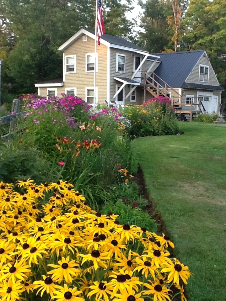 The exterior of a charming two-story building is surrounded by vibrant flower beds filled with yellow and orange blooms. A flagpole stands prominently, displaying the American flag. The building features a pitched roof and stairs leading to a second-level entrance.