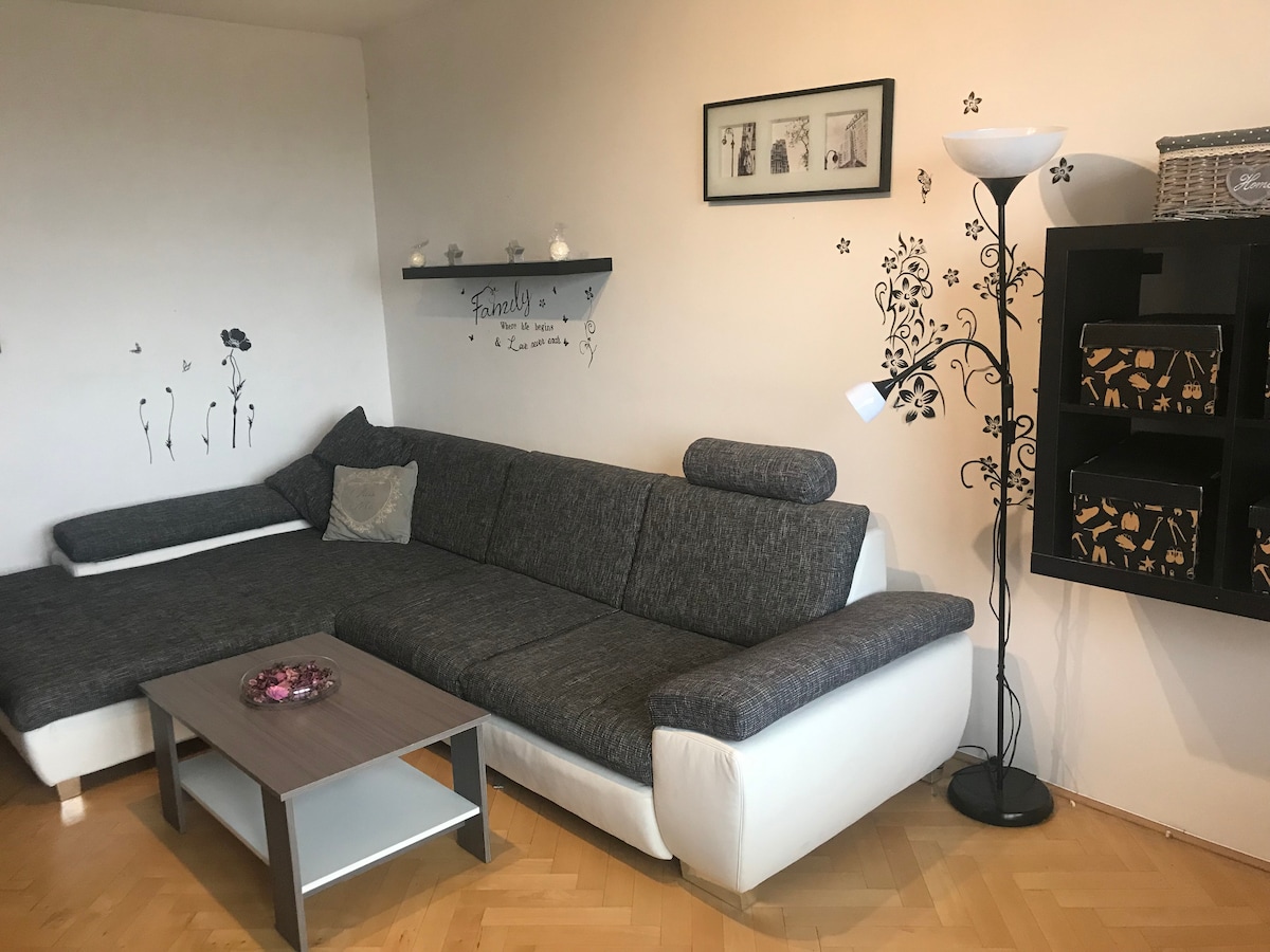 A modern living area is featured, showcasing a gray sectional sofa with white accents. A small coffee table sits in front, adorned with decorative items. A floor lamp provides light, while wall decor adds character. A bookshelf displays elegantly arranged storage boxes.