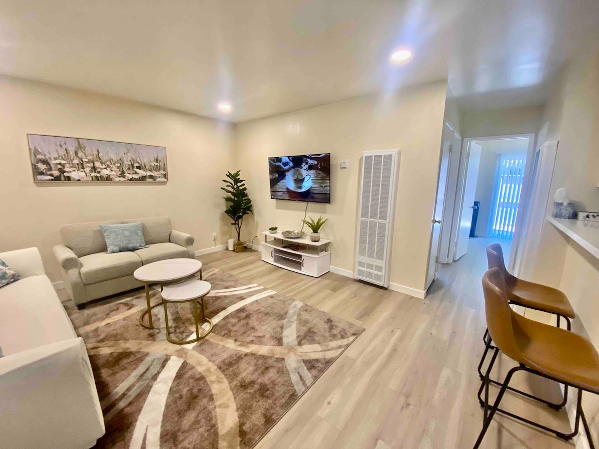 A spacious living area features a cozy sofa and two accent chairs arranged around coffee tables. An entertainment unit displays a wall-mounted TV, while a decorative plant adds a touch of greenery. Soft lighting enhances the inviting ambiance of the room.