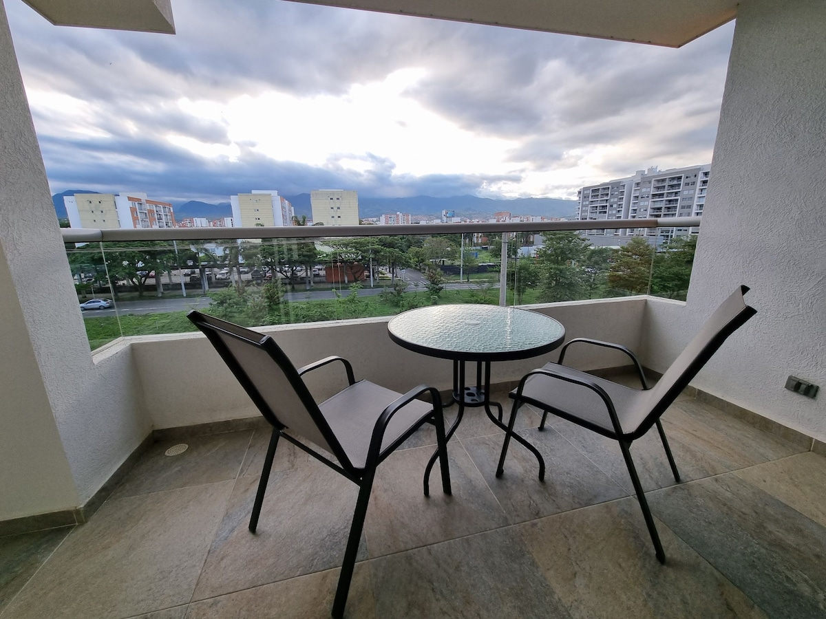 A small balcony features a round glass table accompanied by two black chairs. A panoramic view of the surrounding area is showcased, with greenery and buildings visible in the background under a partly cloudy sky.