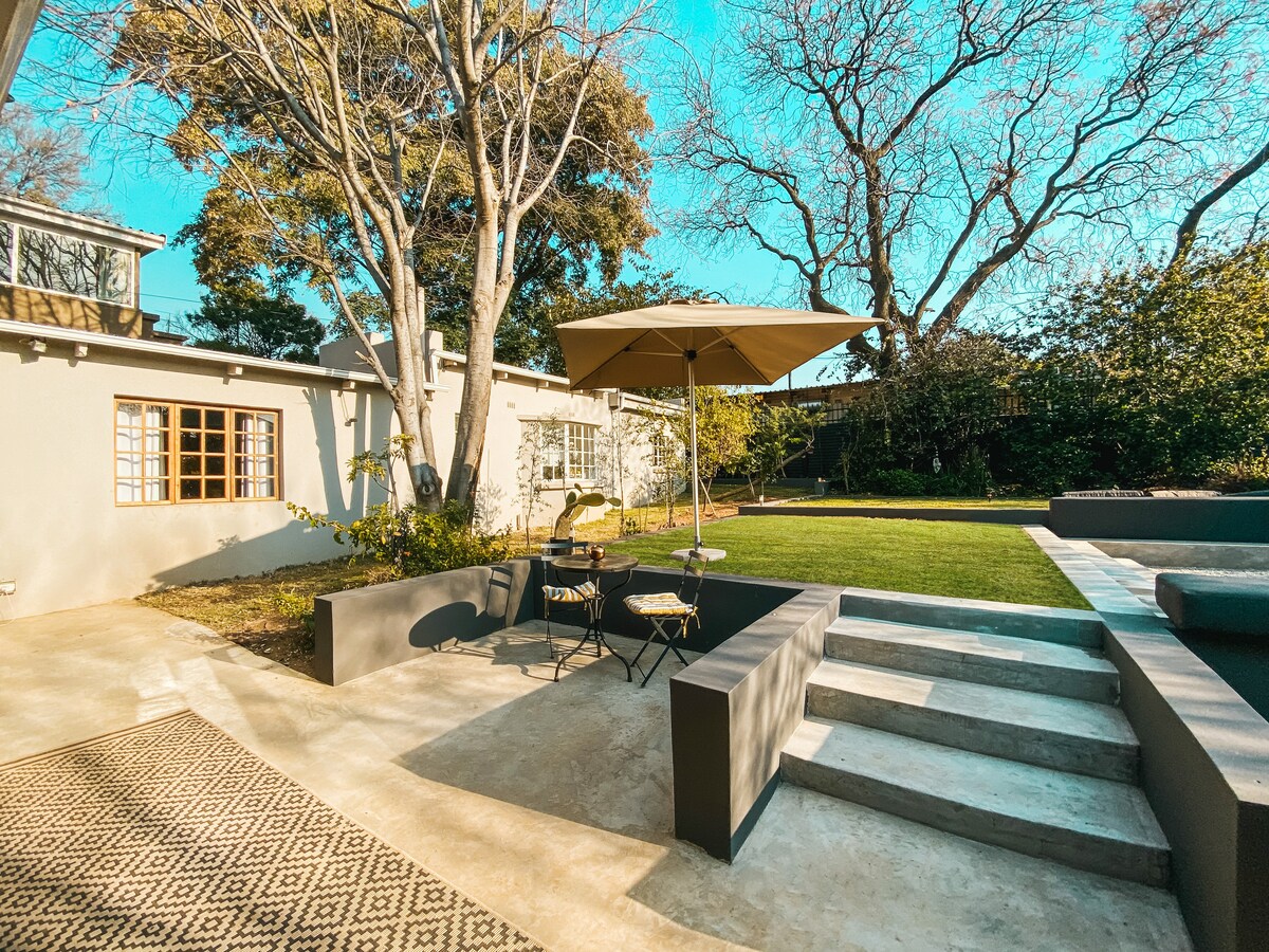 A serene outdoor space features a shaded table with two chairs under a large umbrella. Steps lead to a well-maintained lawn, bordered by trees. Bright sunlight highlights the clean lines of the surrounding architecture, creating an inviting and relaxing atmosphere.