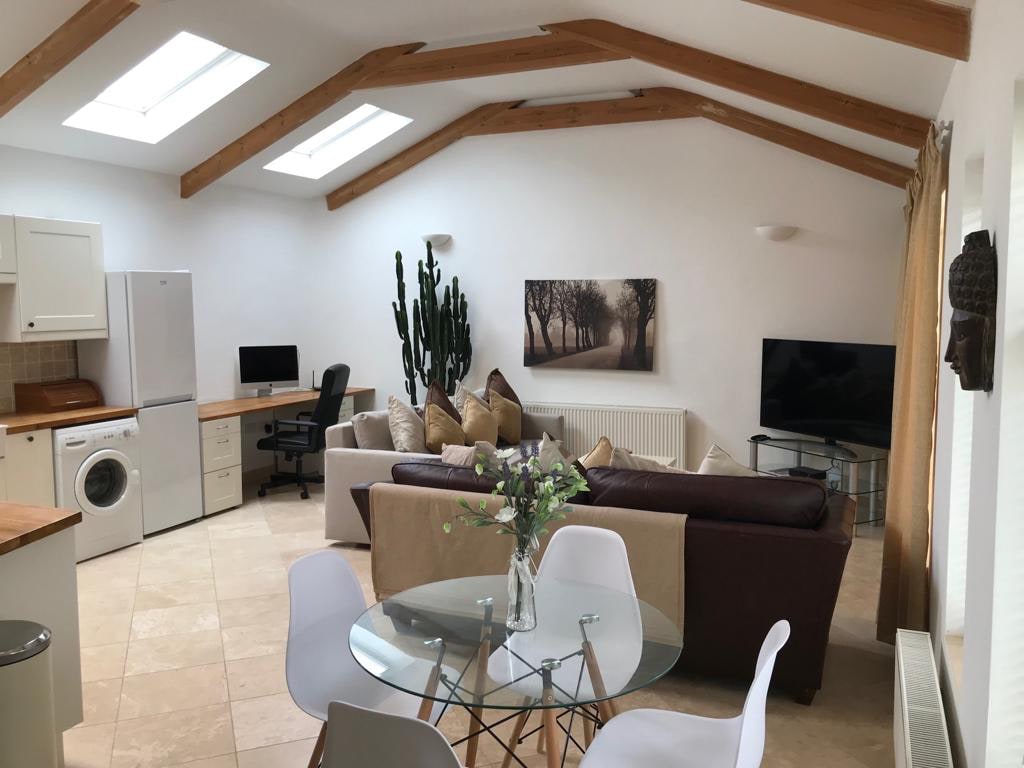 An open plan living area features natural wood beams and three skylights that fill the space with light. A comfortable seating arrangement is positioned around a coffee table. A round dining table accommodates four guests, while a modern kitchen is equipped with essential appliances.